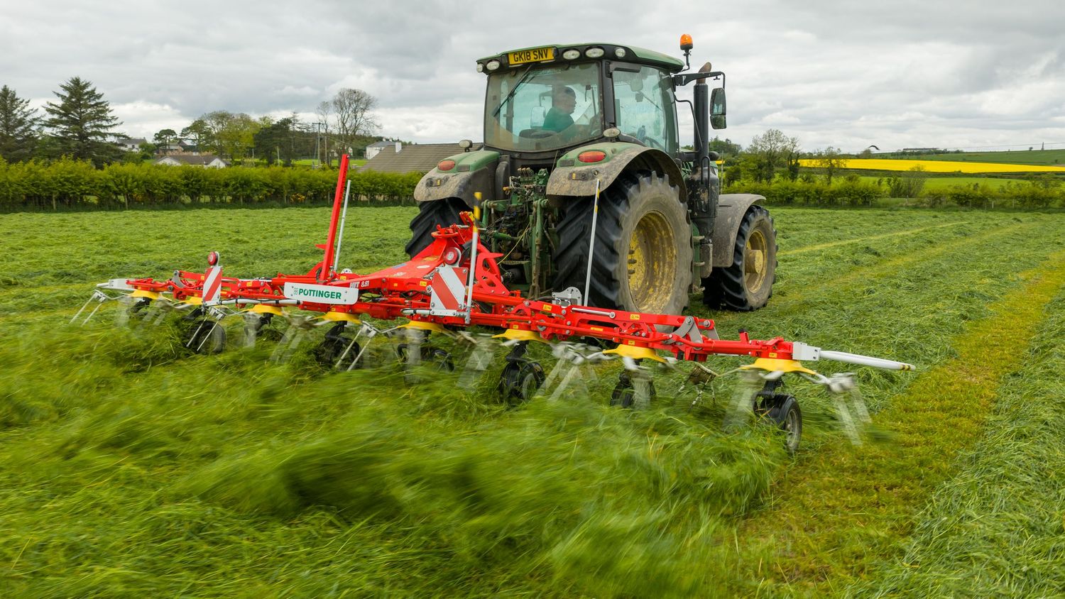 Tractor pulling a red multi-rotor rake through a green field, creating neat rows of cut grass.