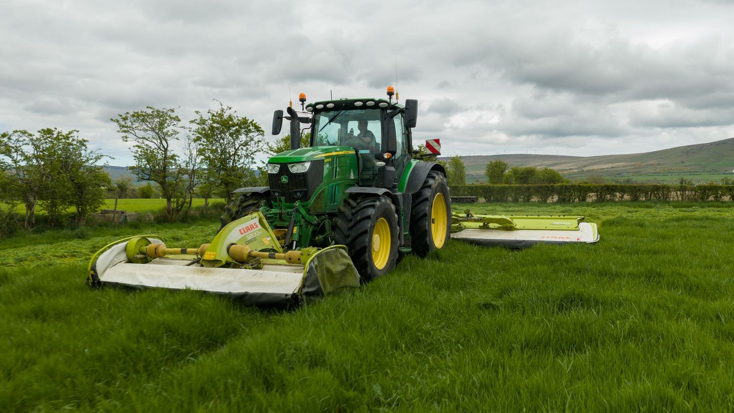 John Deere green tractor with dual mowers cutting grass in a field on an overcast day.