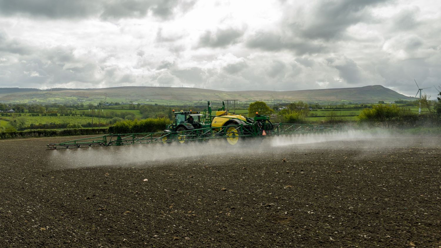A yellow tractor spraying pesticides on a large dark soil farm field with rolling green hills under cloudy skies.