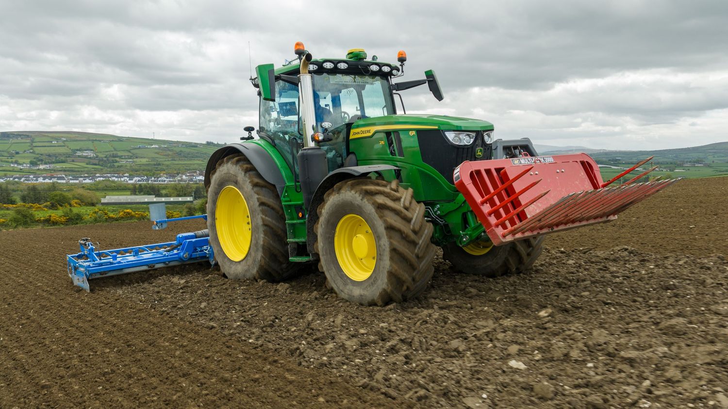 Green John Deere tractor plowing a field with a red attachment on overcast day in rural landscape.