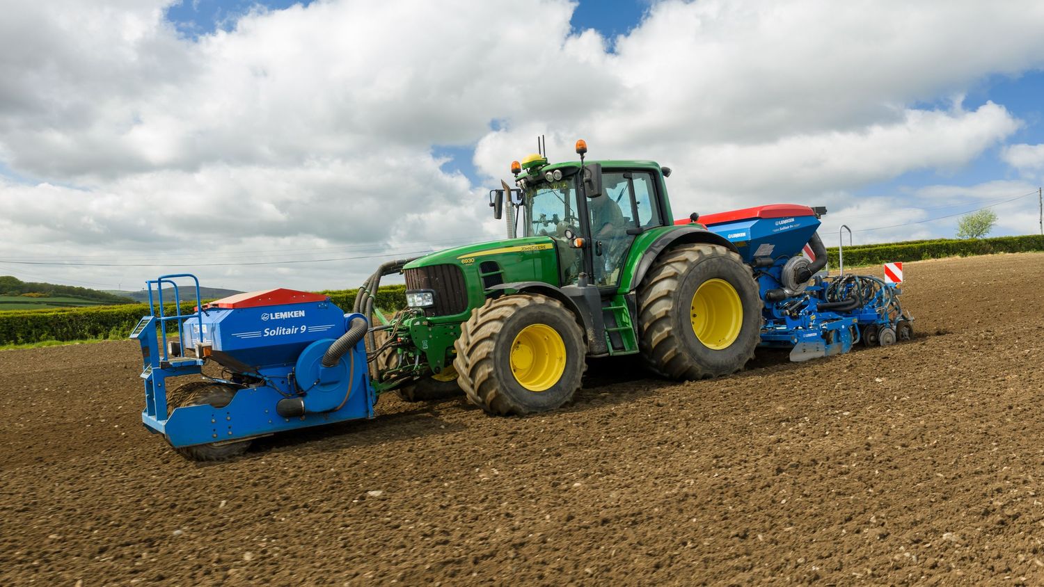 Green John Deere tractor with blue seeder attachment plowing a field under cloudy skies.