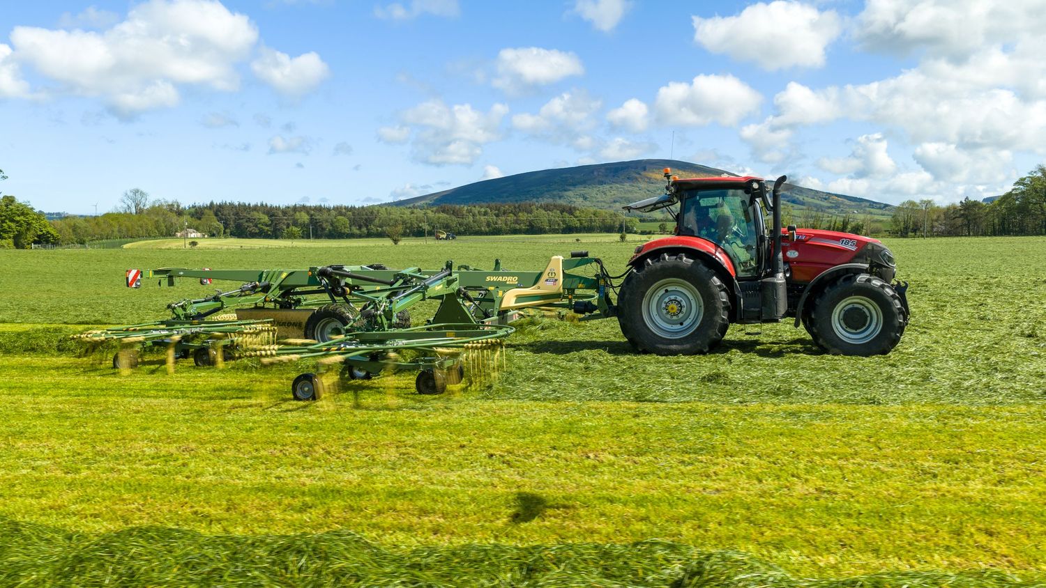 Red tractor with green hay rake attachment working in a lush green field on a sunny day.