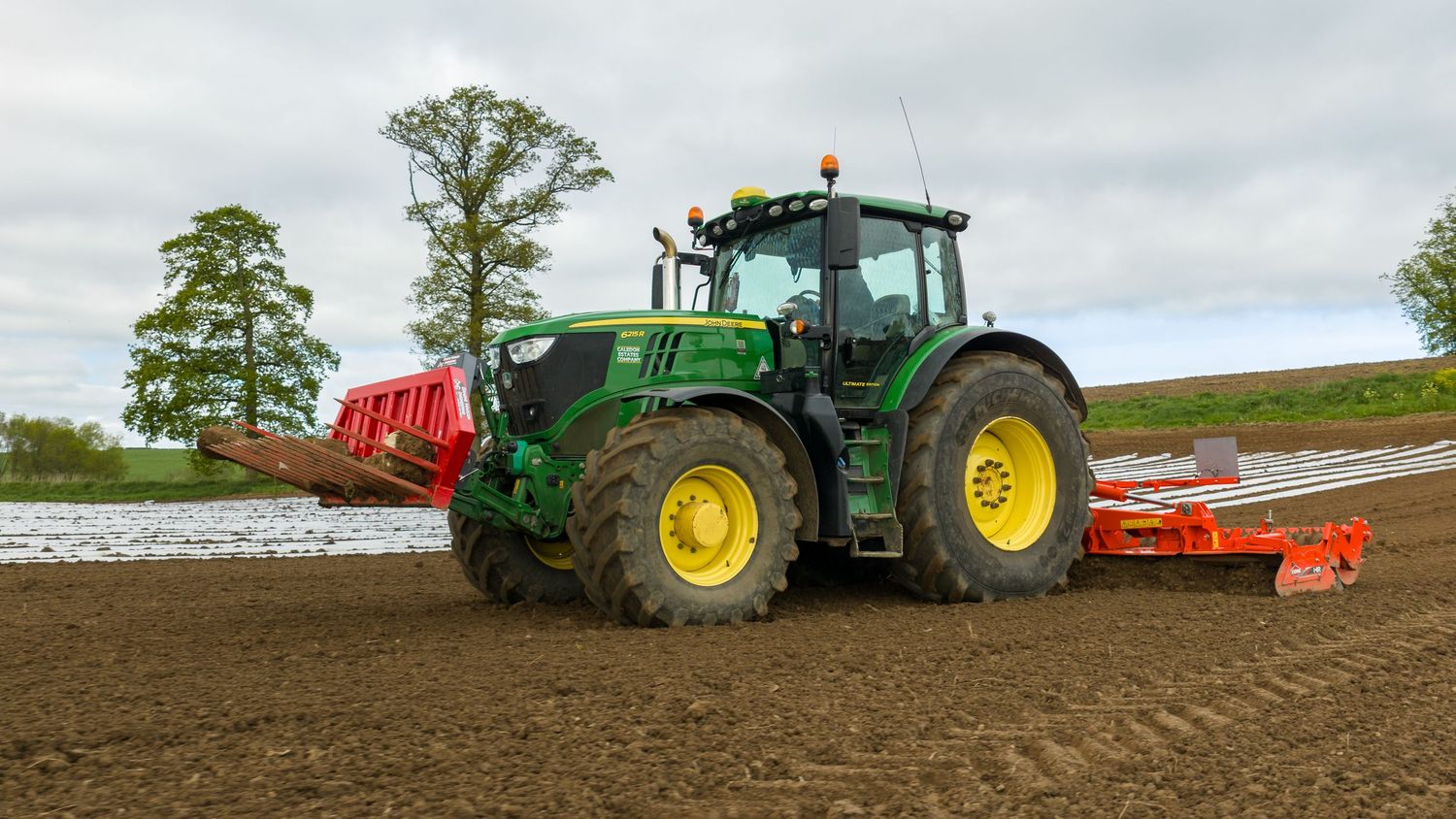 Green John Deere tractor with red seeding equipment cultivating a large agricultural field in spring.