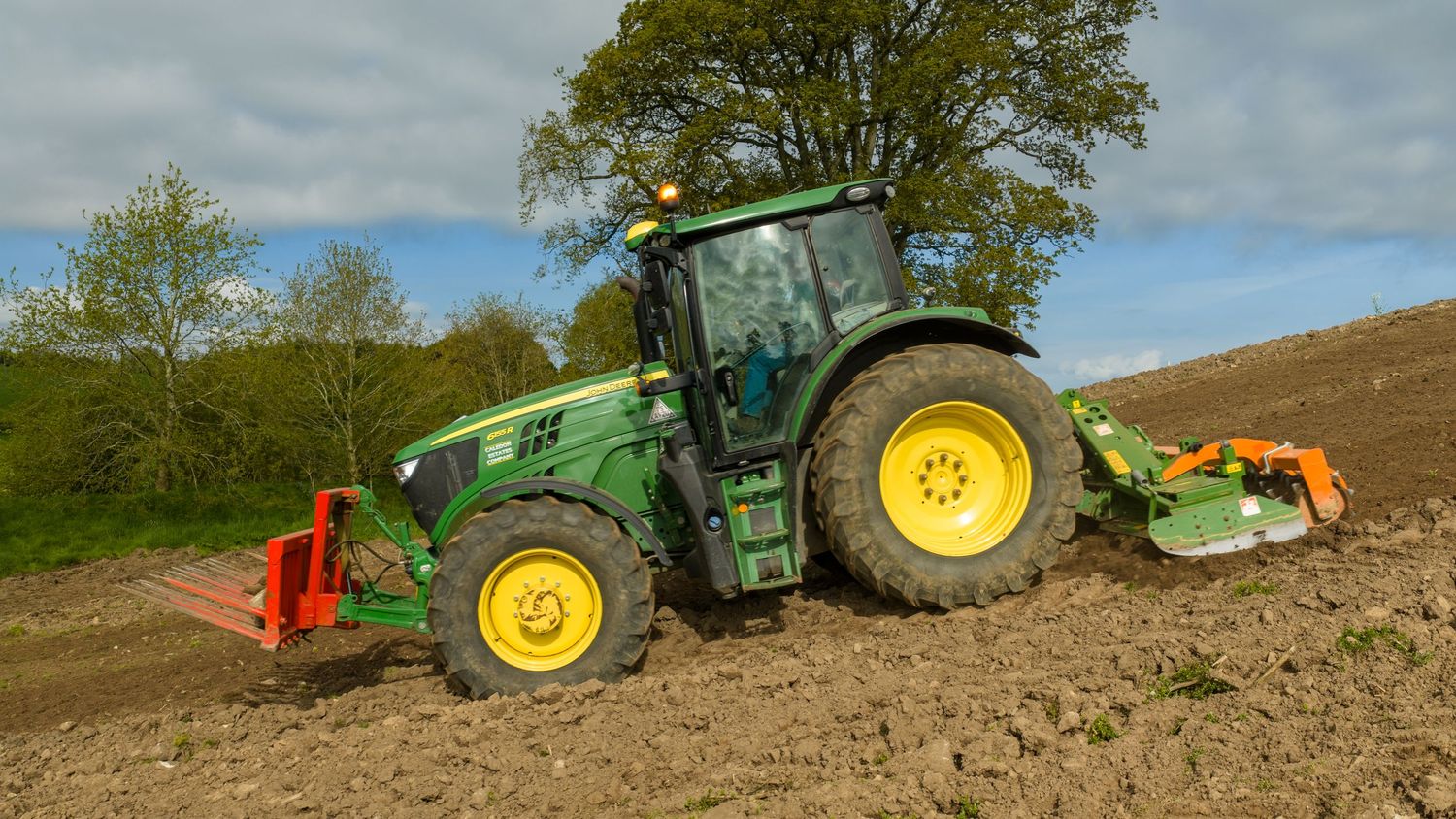 Green John Deere tractor plowing a field with attached tillers on a sunny day near trees.