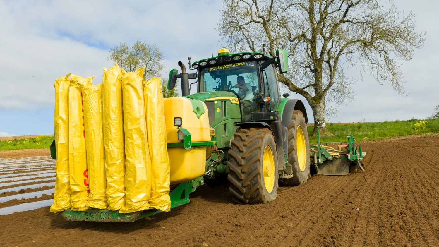 Green John Deere tractor with yellow equipment attachment tilling dark soil in agricultural field.