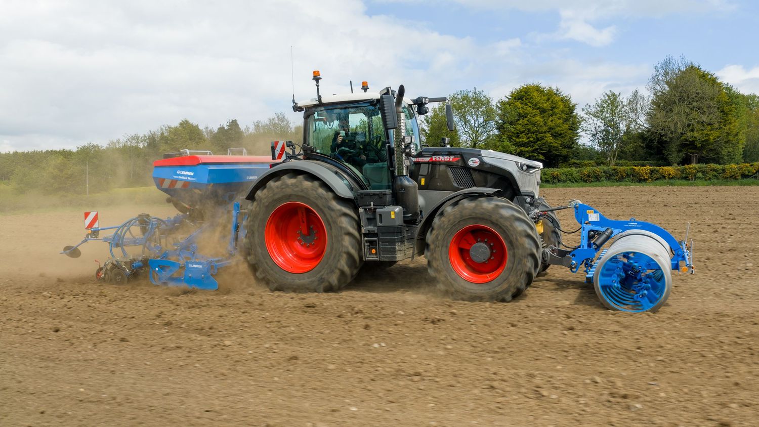 White and blue tractor with red wheels plowing dusty farmland field on a sunny day.