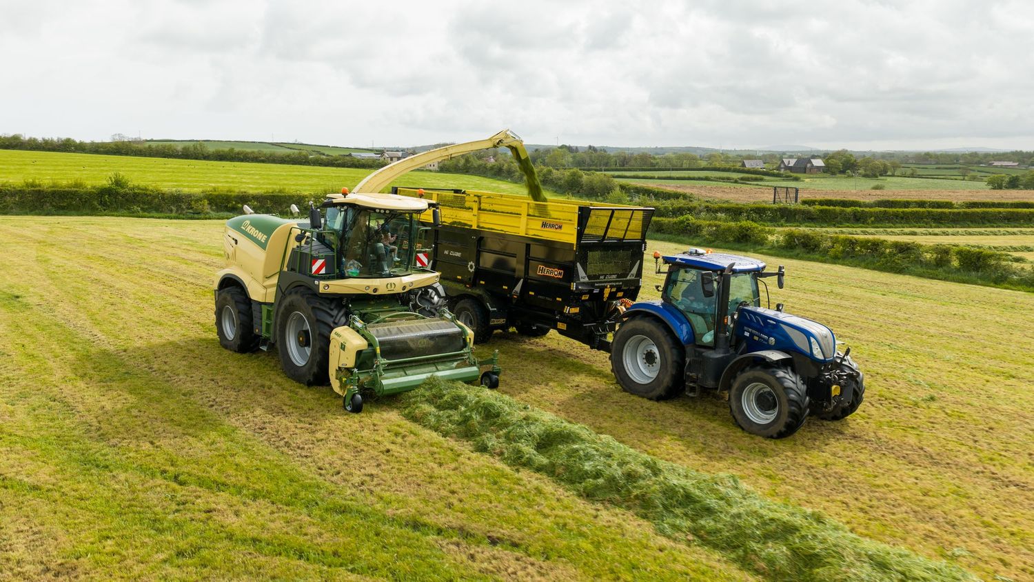 A yellow Krone forage harvester and blue New Holland tractor harvesting silage in a green field.