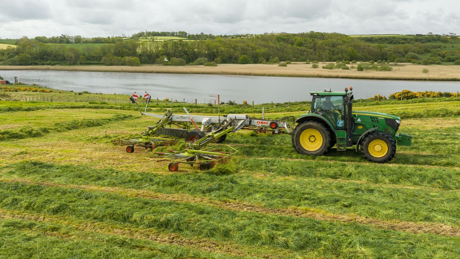 Green John Deere tractor harvesting grass in a scenic field beside a river on a cloudy day.