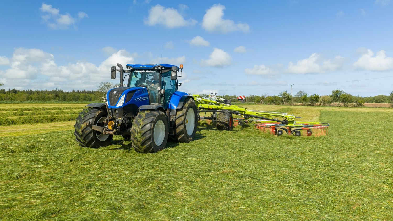 Blue New Holland tractor pulling green Claas rake implement across lush green field under blue sky.