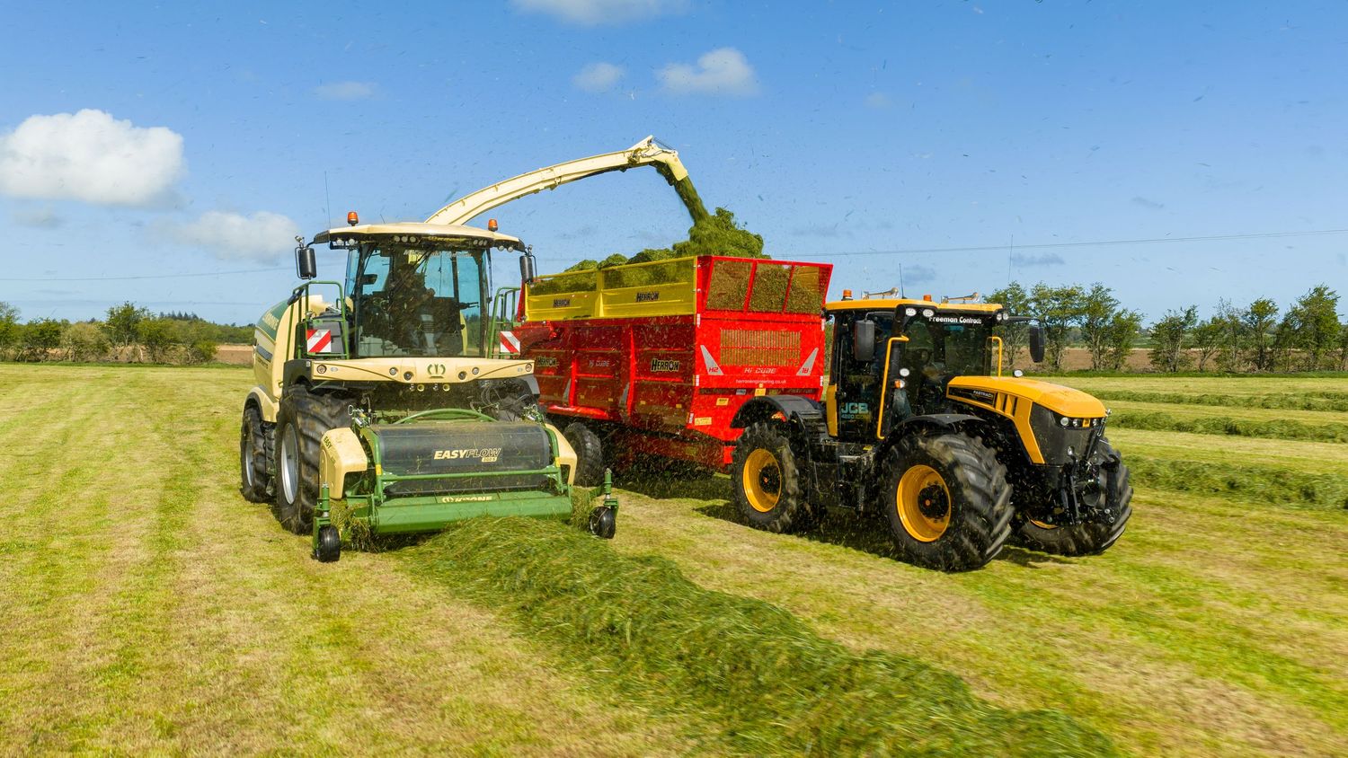 Yellow forage harvester unloading silage into red trailer pulled by yellow tractor in green field.