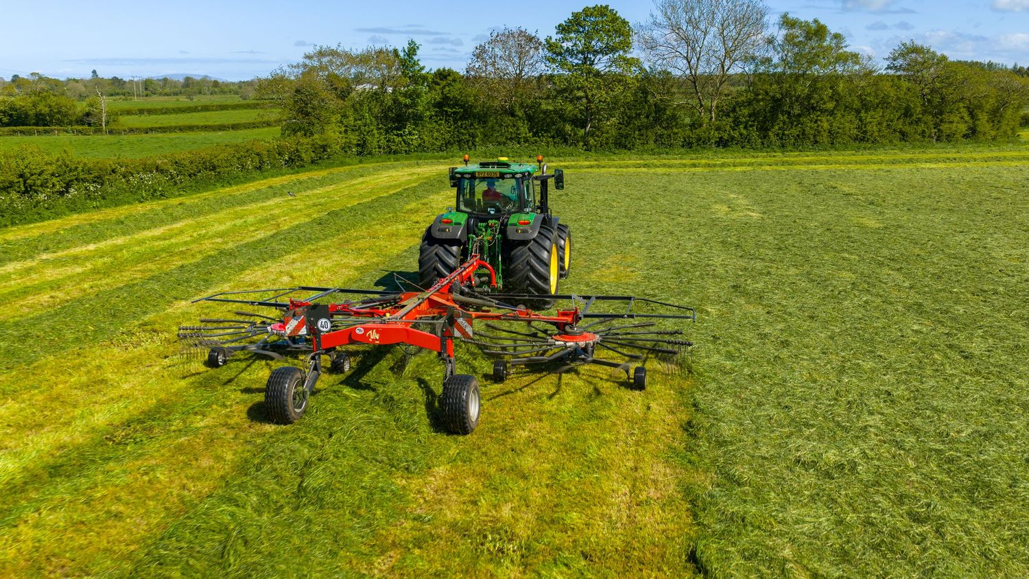 Green tractor with red tedder attachment spreading cut grass in a lush green field on a sunny day.