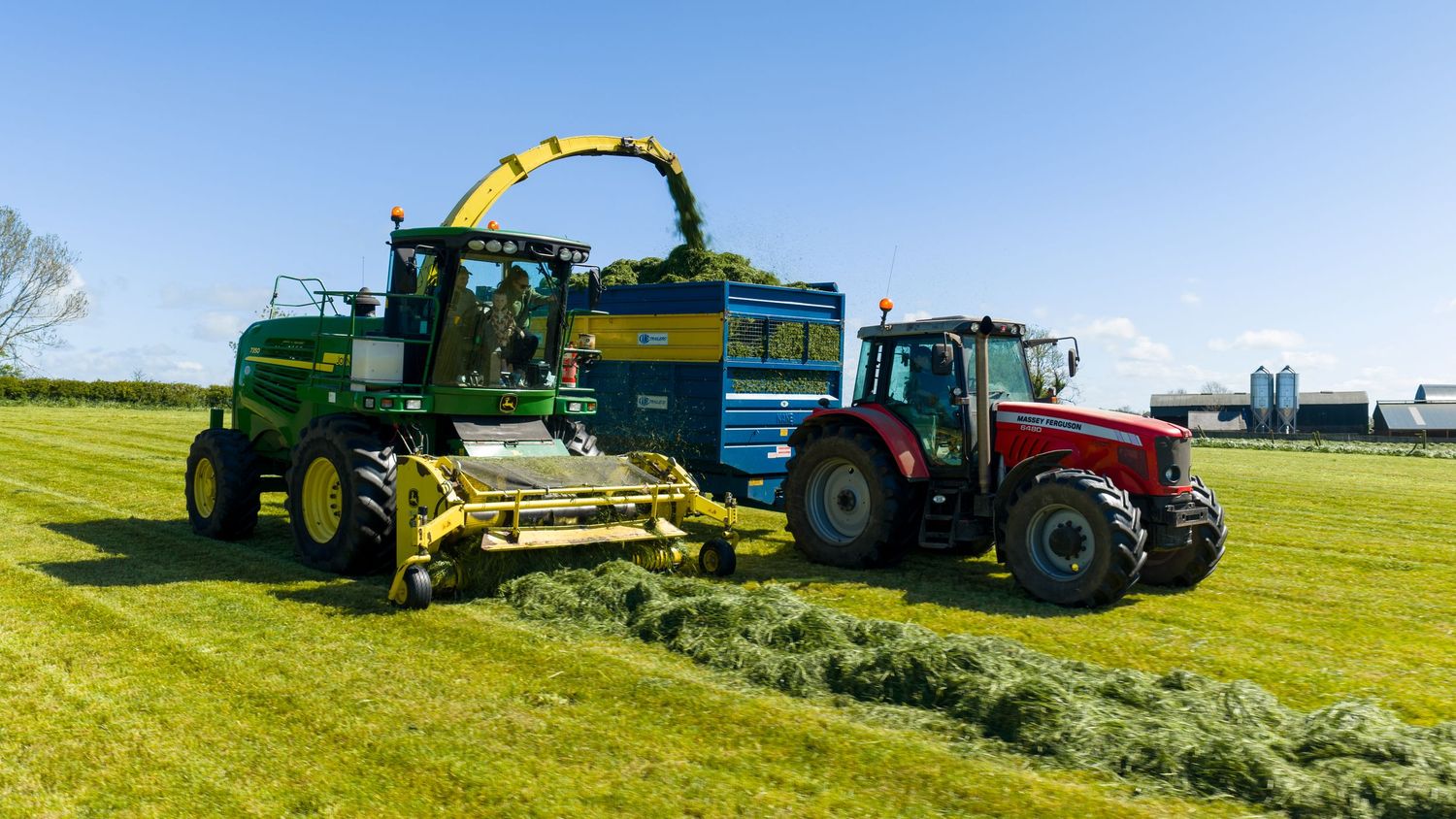 Green John Deere forage harvester working alongside red tractor in lush field on sunny day.