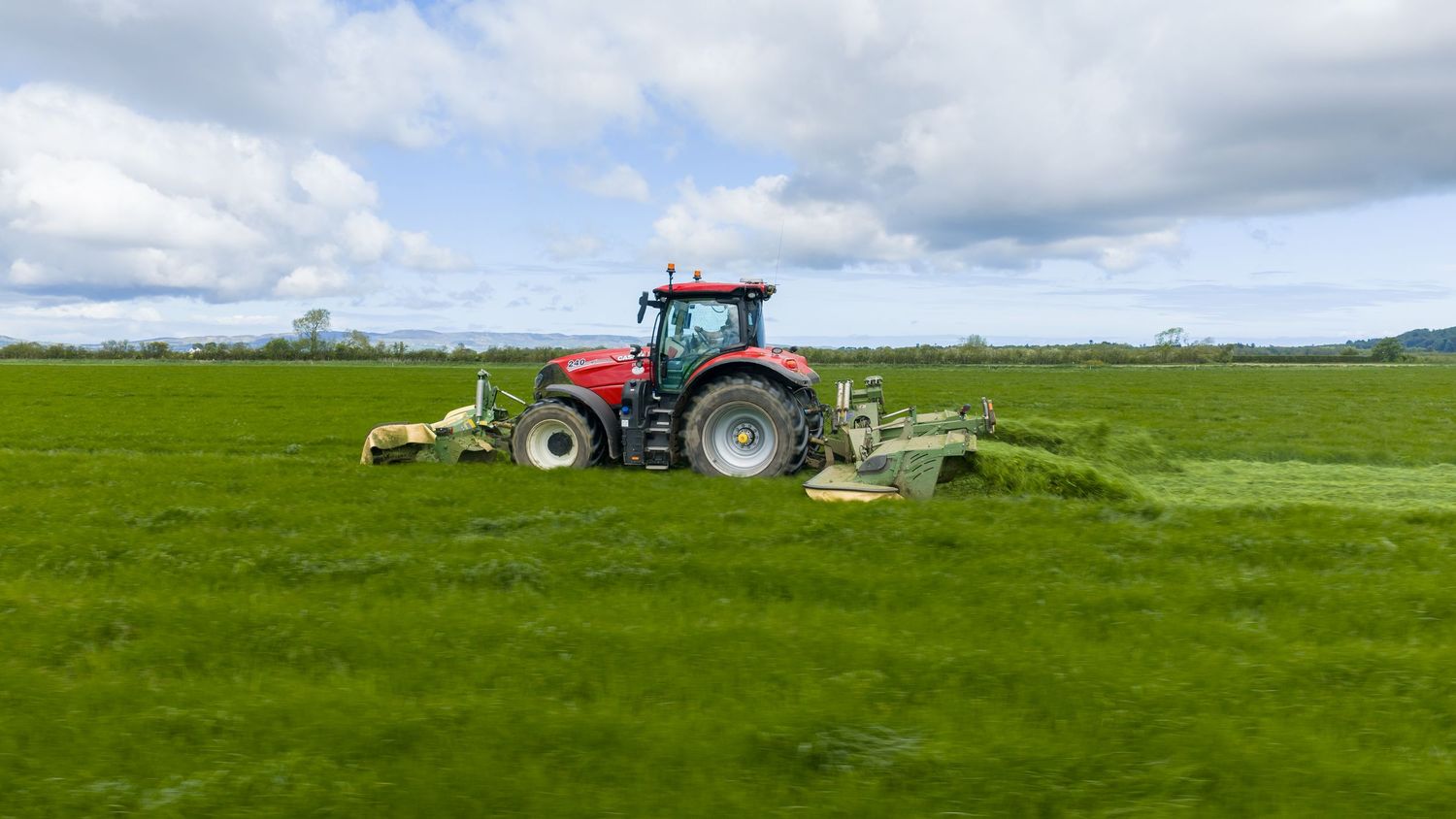 Red tractor mowing lush green grass field on a sunny day with blue skies and white clouds in background.