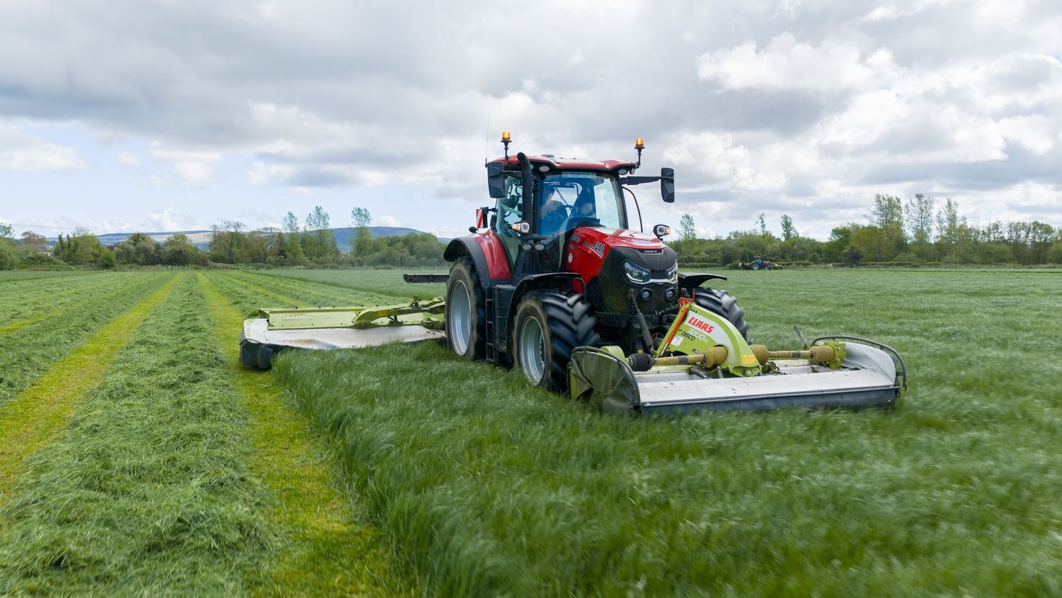 Red tractor with wide mower attachment cutting green grass in a field on a cloudy day.