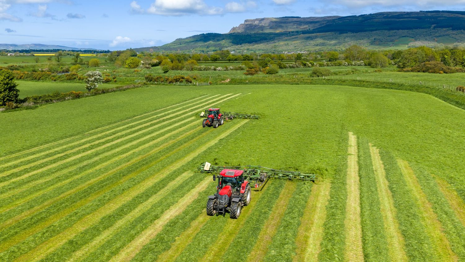 Aerial view of two red tractors mowing green fields in a scenic rural landscape with hills in the background.