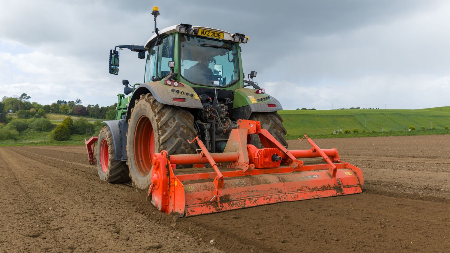 Green Fendt tractor with orange rotary tiller attachment cultivating a field under cloudy skies.