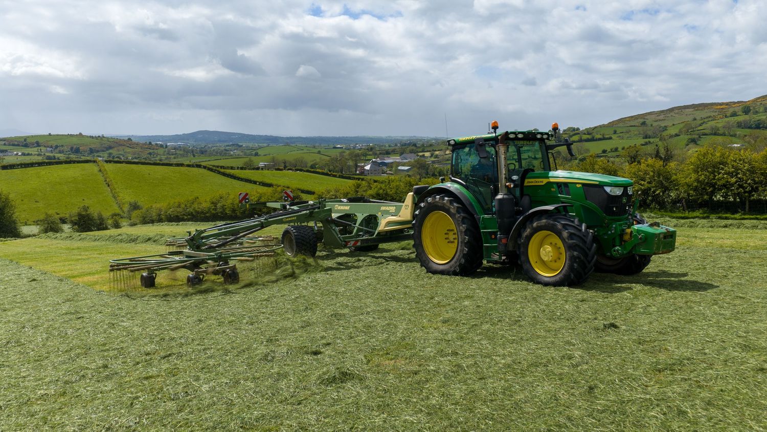 Green John Deere tractor with hay rake attachment working in a lush green field with rolling hills in background.