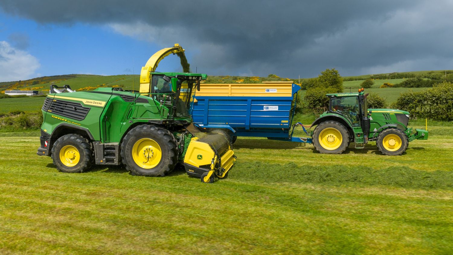 John Deere green forage harvester and tractor with blue trailer working in a lush green field under cloudy skies.