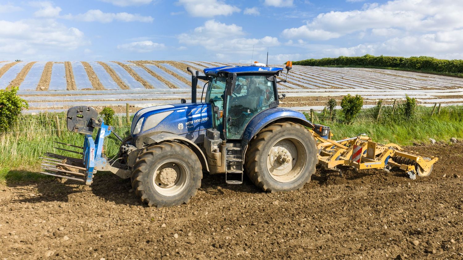 Blue New Holland tractor tilling soil in a farm field with greenhouse structures visible in the background.
