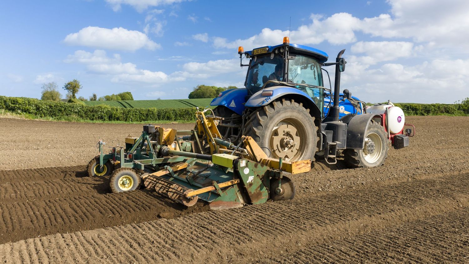 Blue New Holland tractor with farming equipment tilling a brown agricultural field on a sunny day.