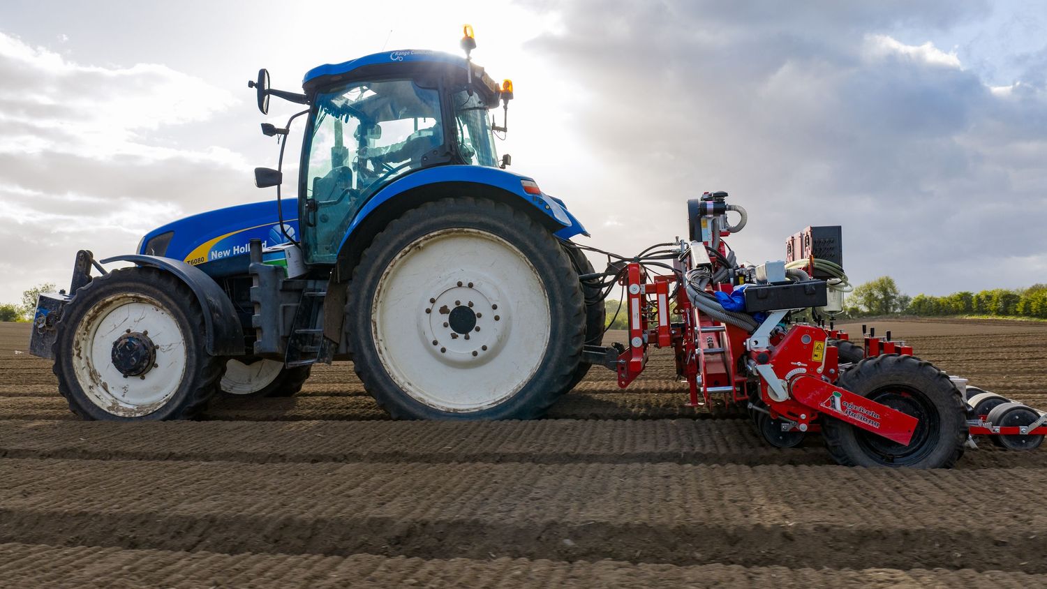 Blue New Holland tractor with red precision seeding equipment working in a farm field under dramatic cloudy skies.