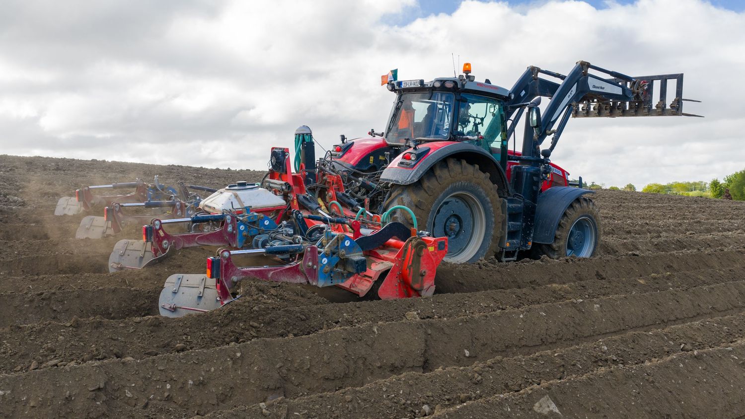 Red tractor with front loader plowing and seeding a large agricultural field under cloudy skies.
