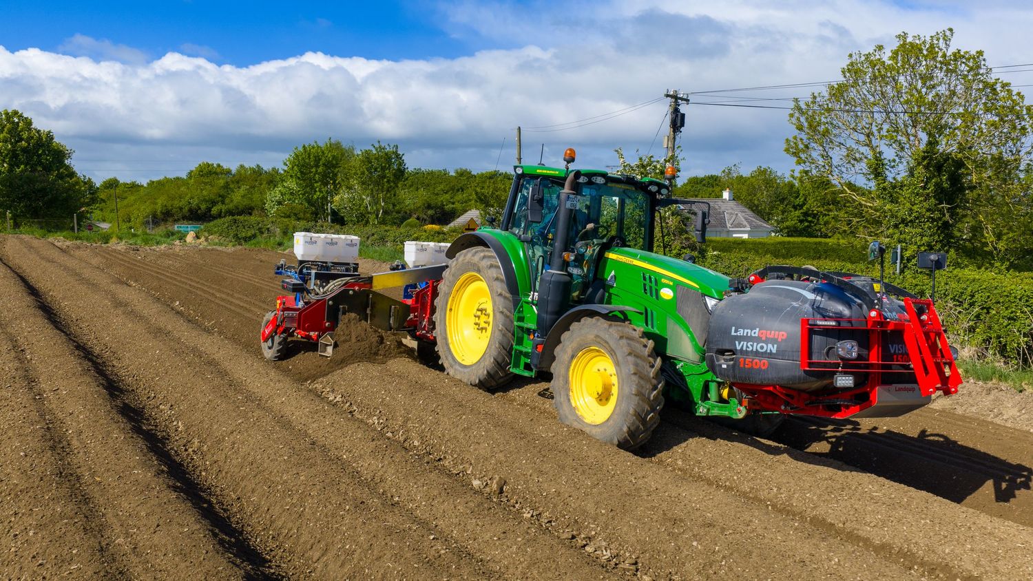 Green John Deere tractor with red planting equipment working rows in a freshly tilled agricultural field.