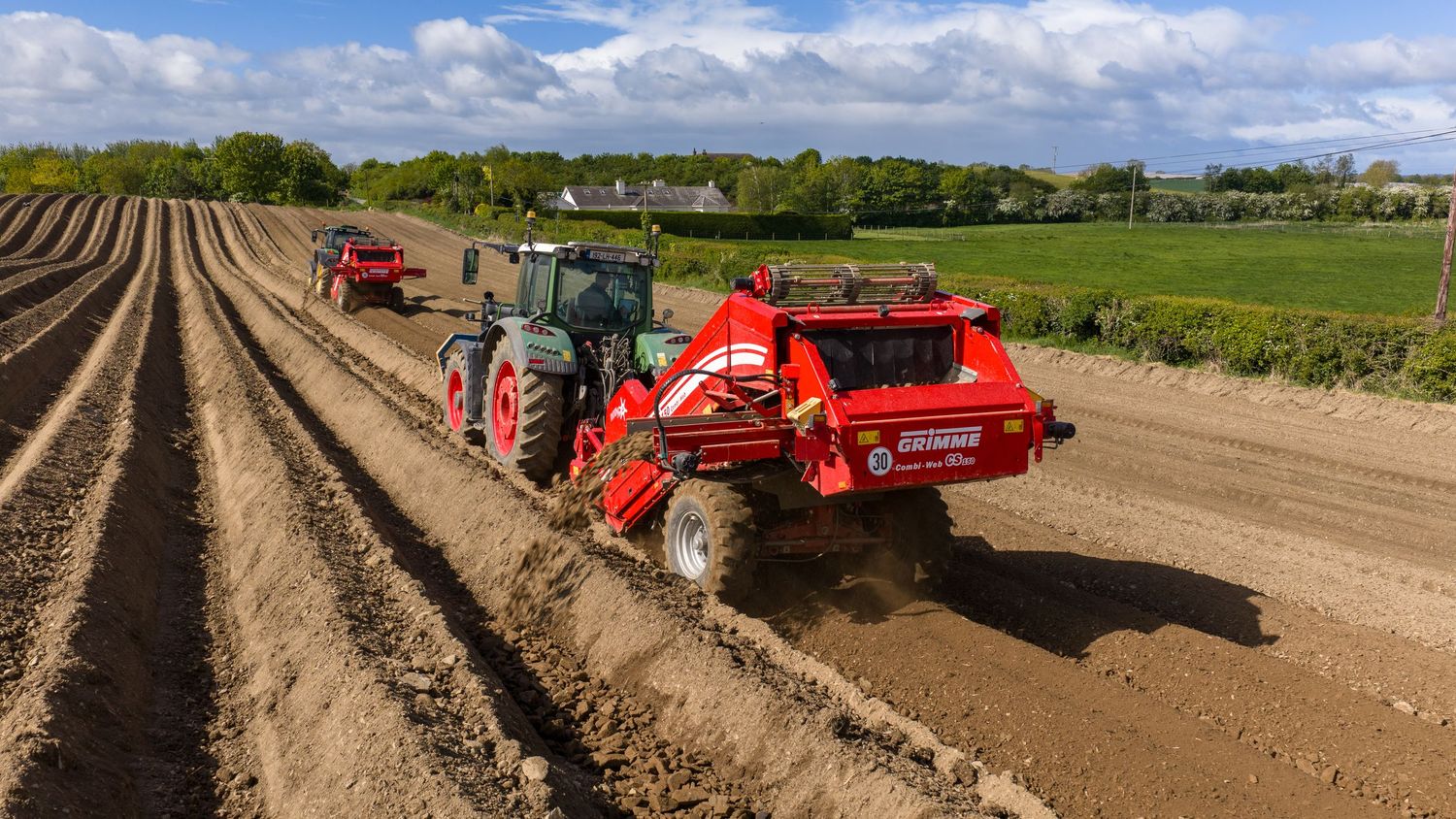 Red tractor planting potatoes in rows on a large farm field under a partly cloudy sky.