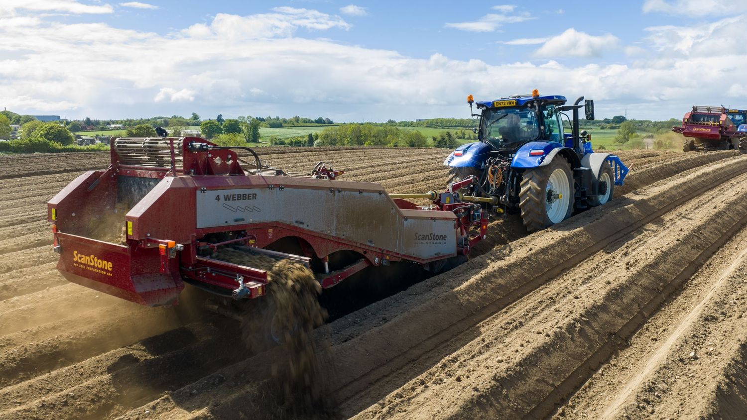 Blue tractor pulling red Grimme potato harvester equipment across freshly plowed agricultural field rows.