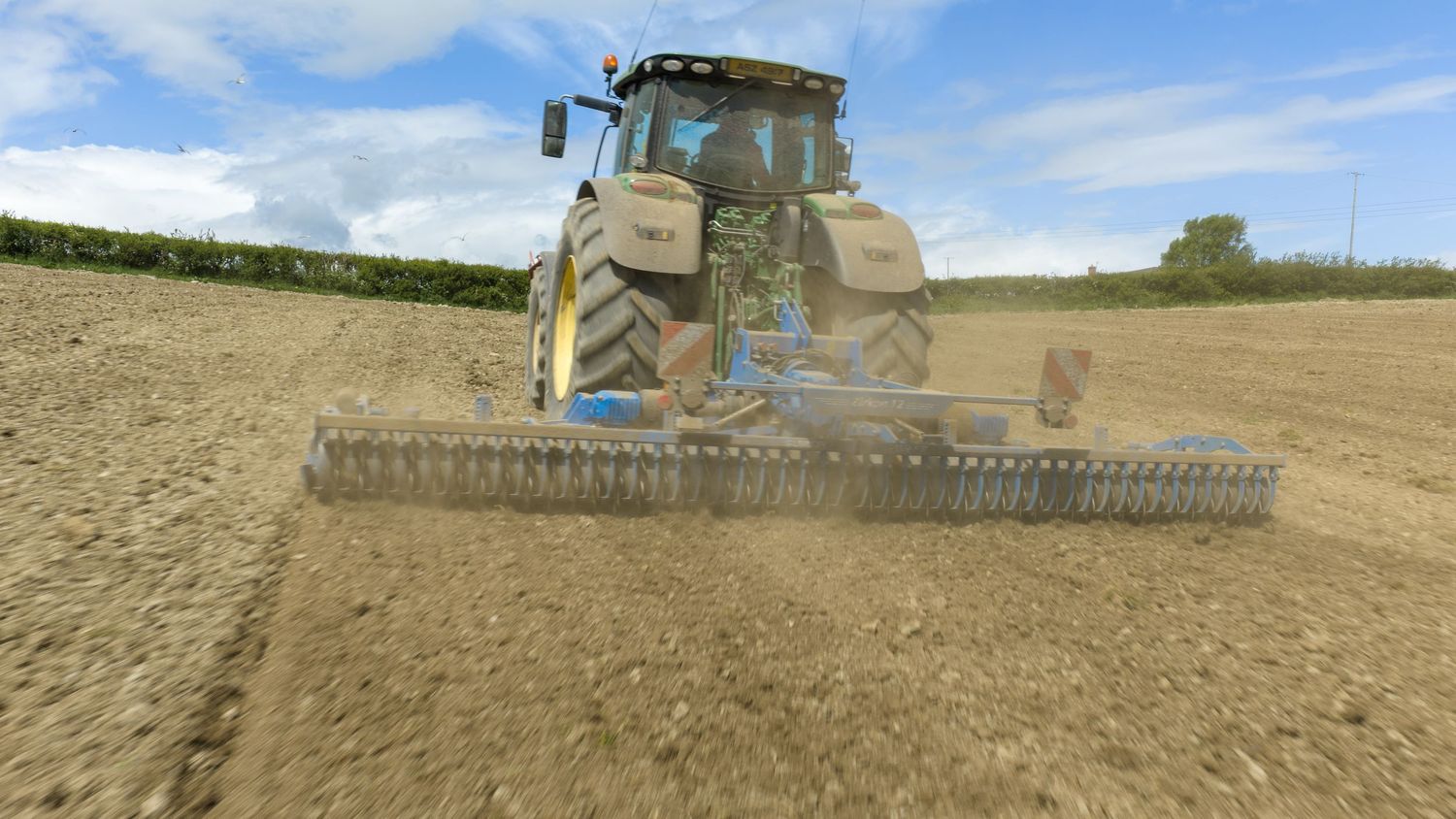 Yellow tractor pulling a soil cultivator across a plowed agricultural field under a blue sky.