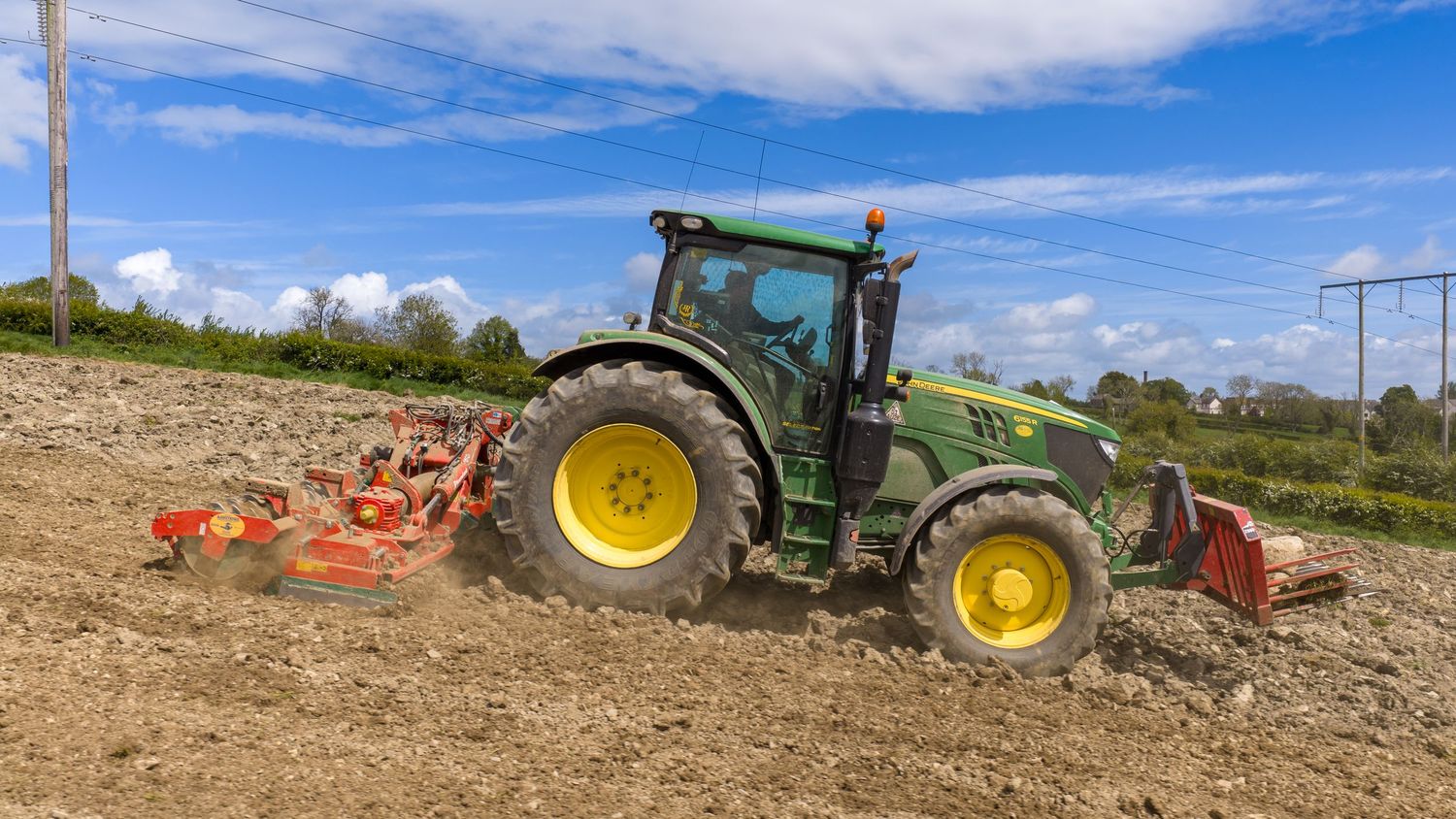 Green John Deere tractor with yellow wheels plowing a field under a blue sky with white clouds.