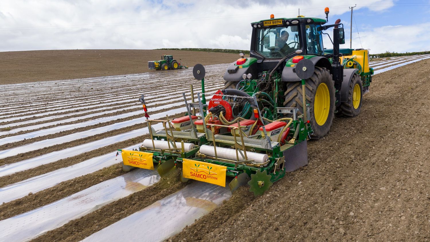 Green John Deere tractor with yellow planting equipment laying plastic mulch film on agricultural field rows.