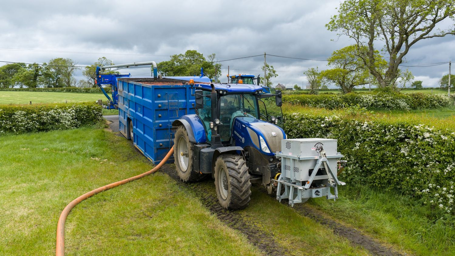 Blue tractor with trailer connected to orange hose pipe in rural countryside field setting.