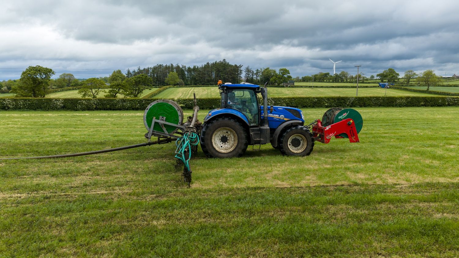 Blue tractor with slurry spreading equipment working in a lush green field on a cloudy day.