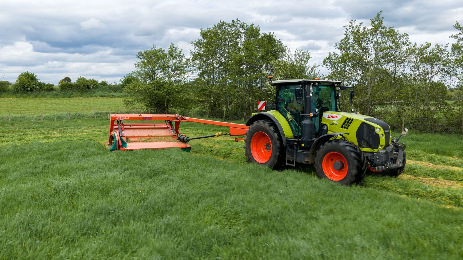 Green CLAAS tractor pulling a red mower attachment across a lush green field under a cloudy sky.