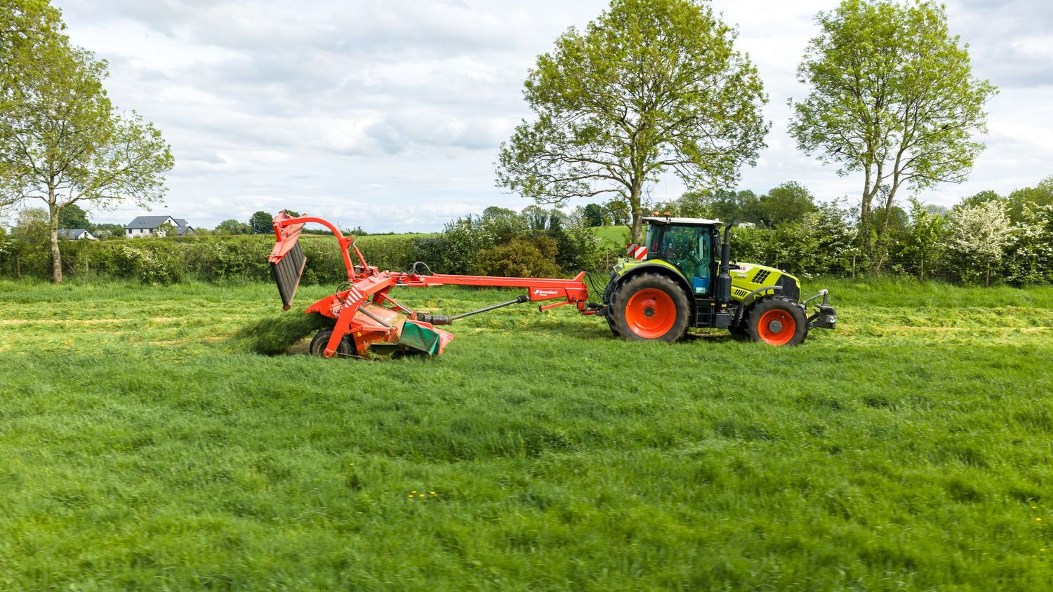 Green tractor with red mower attachment cutting lush grass in an open field surrounded by trees.