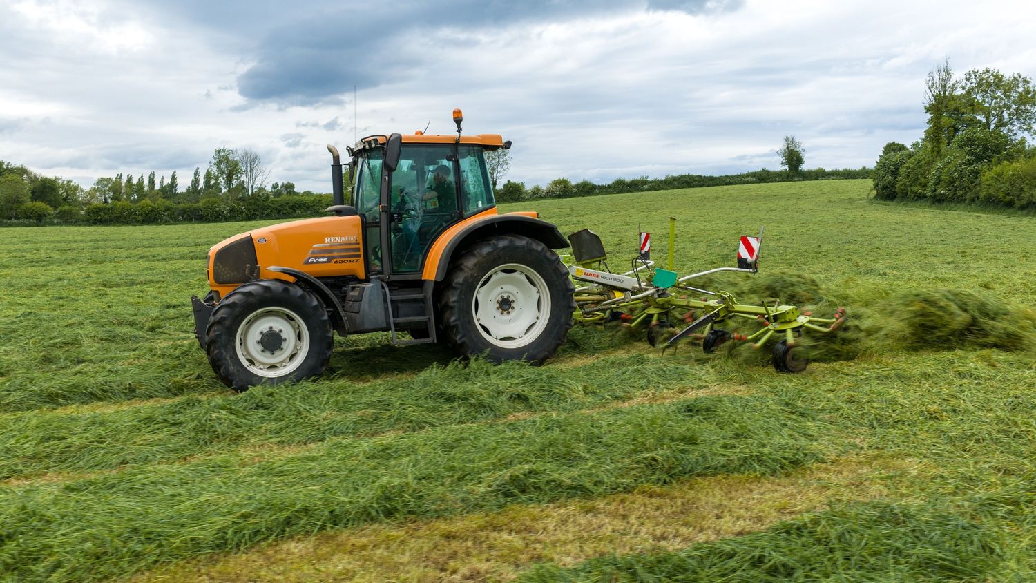 Orange tractor mowing green grass field with hay rake attachment on a cloudy day in a rural countryside setting.