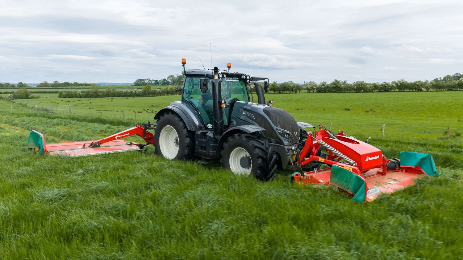 A black Valtra tractor with red mowers cutting green grass in a lush farm field under a cloudy sky.