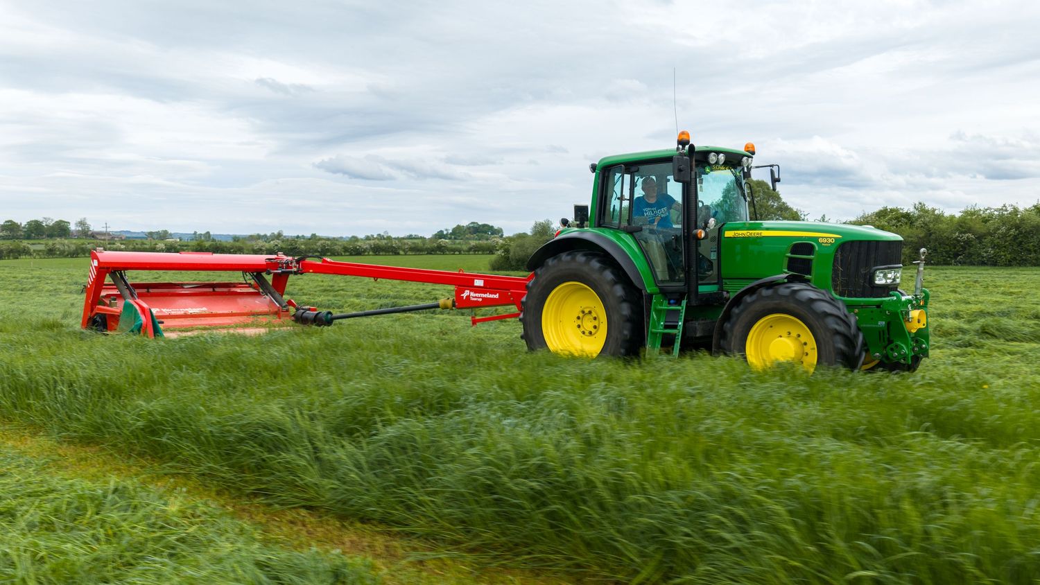 Green John Deere tractor mowing lush green grass field with red mower attachment on a cloudy day.