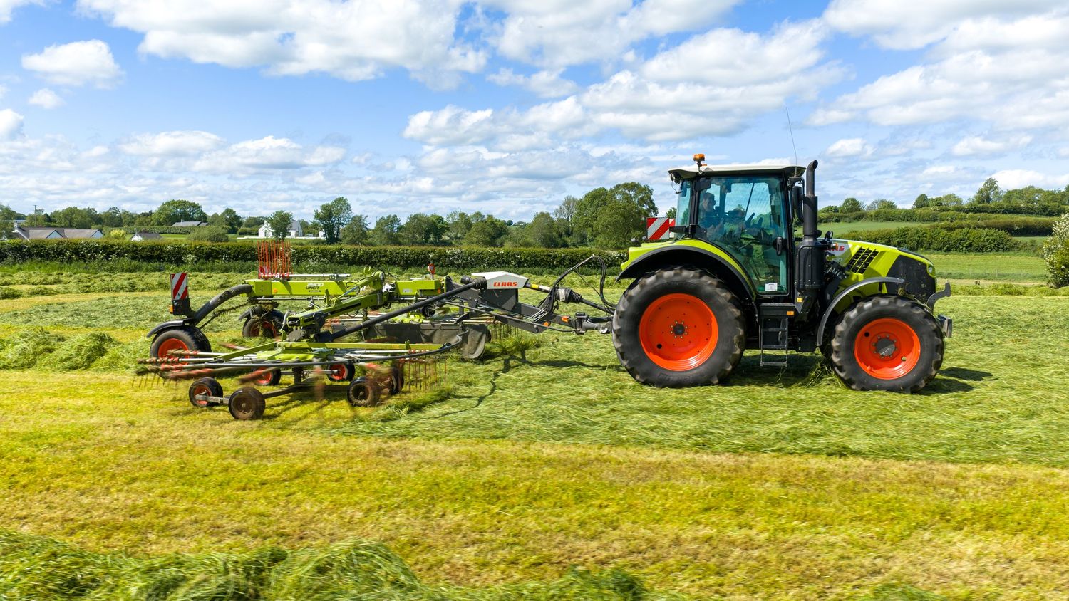 Green CLAAS tractor pulling hay rake equipment across lush farmland under a bright blue sky with white clouds.