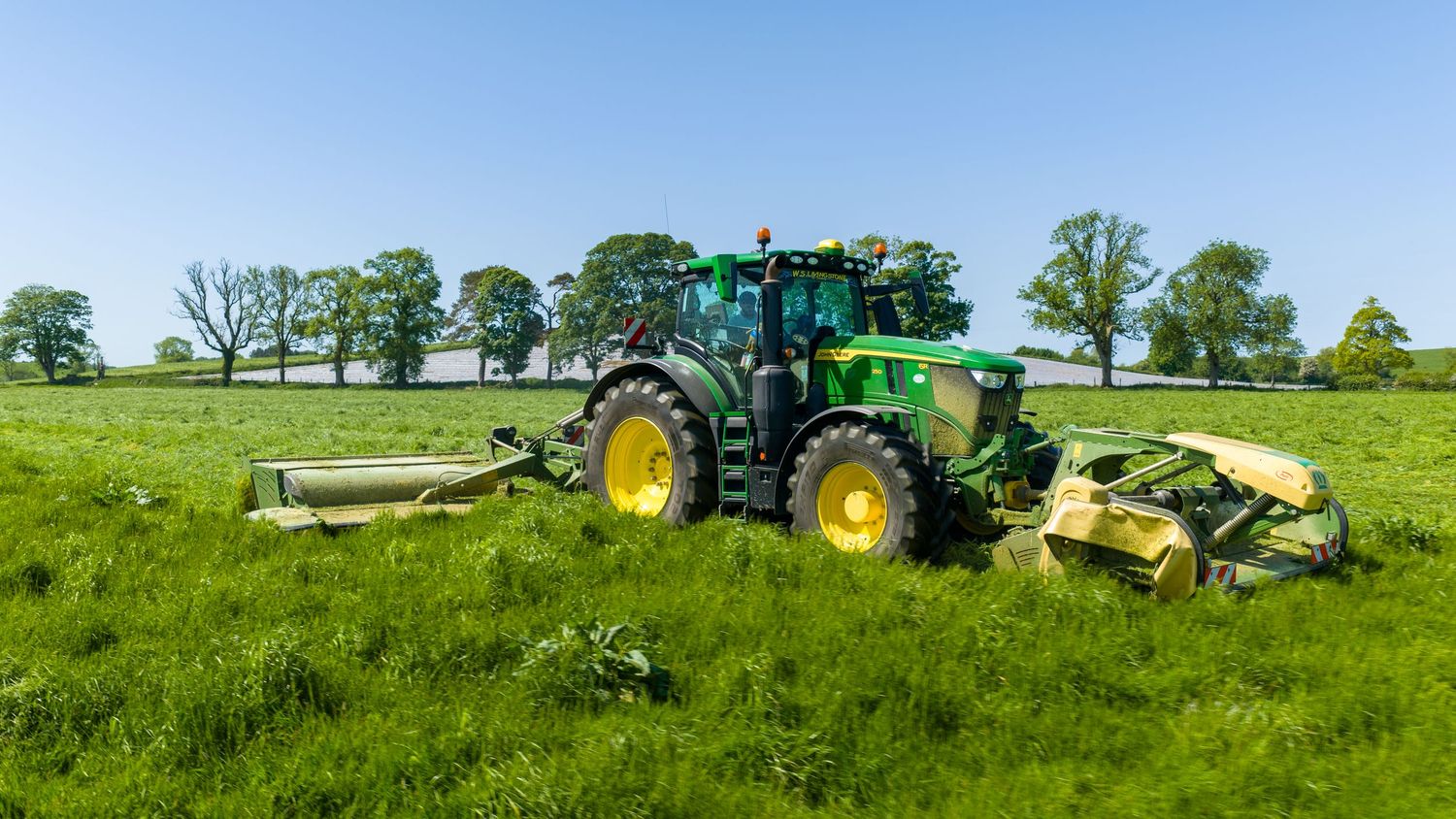 Green John Deere tractor mowing a lush green field on a sunny day with trees in the background.