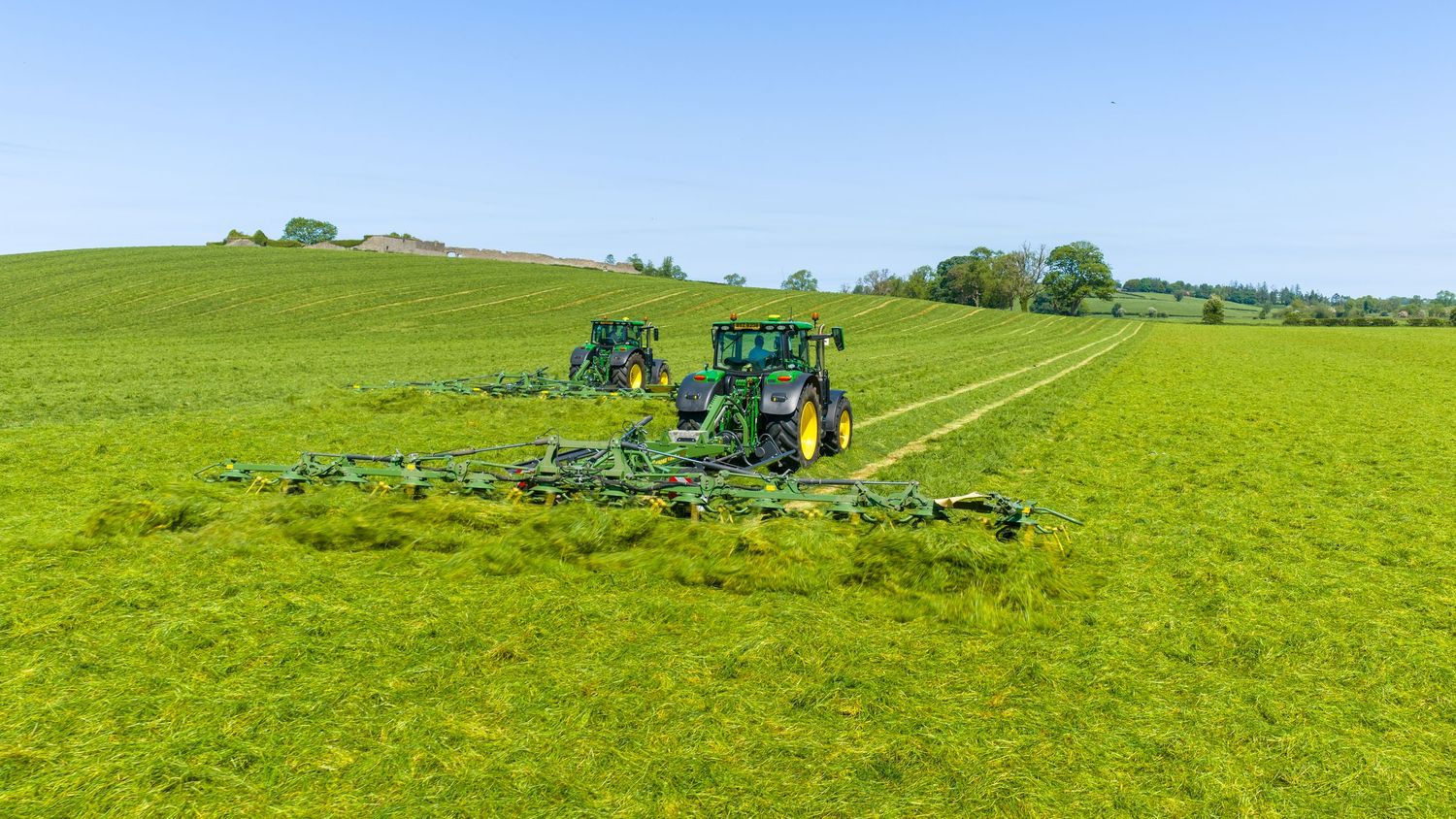 Two green John Deere tractors operating large hay rakes on a lush green hillside field under a clear blue sky.