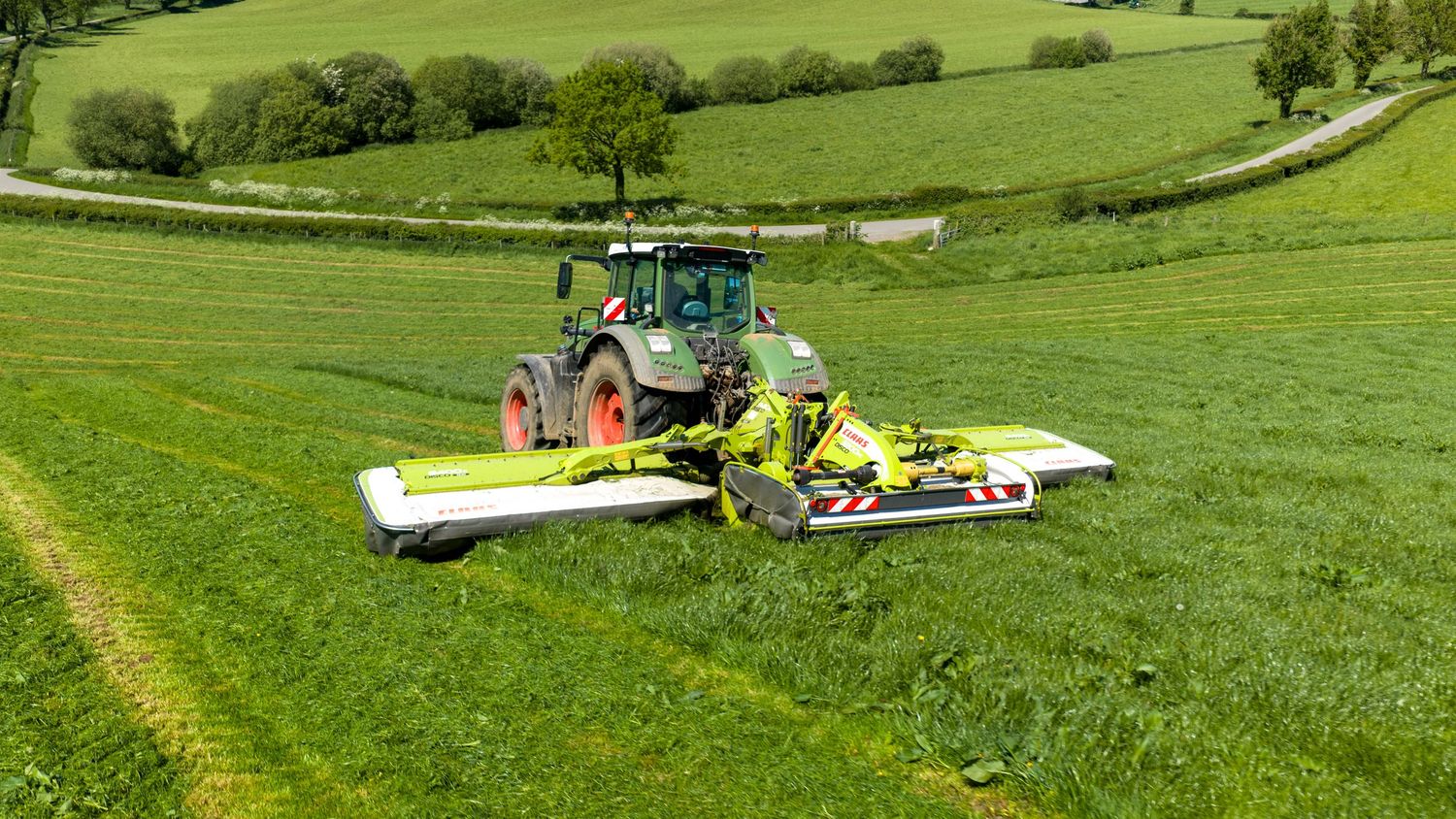 Green Fendt tractor with yellow mower attachment cutting lush grass in a scenic countryside field.