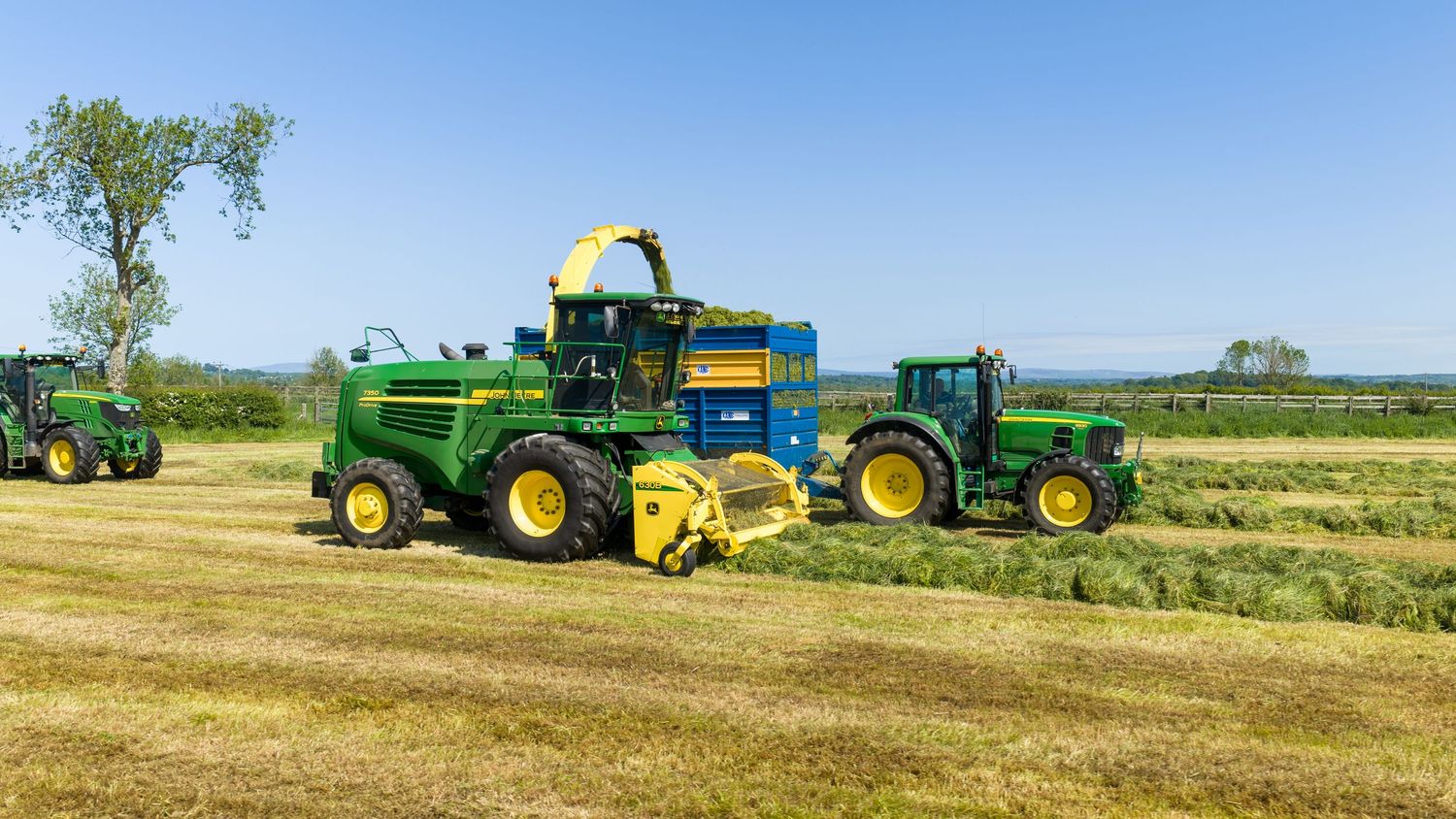 John Deere green forage harvester and tractors working in a sunny field during hay harvesting season.
