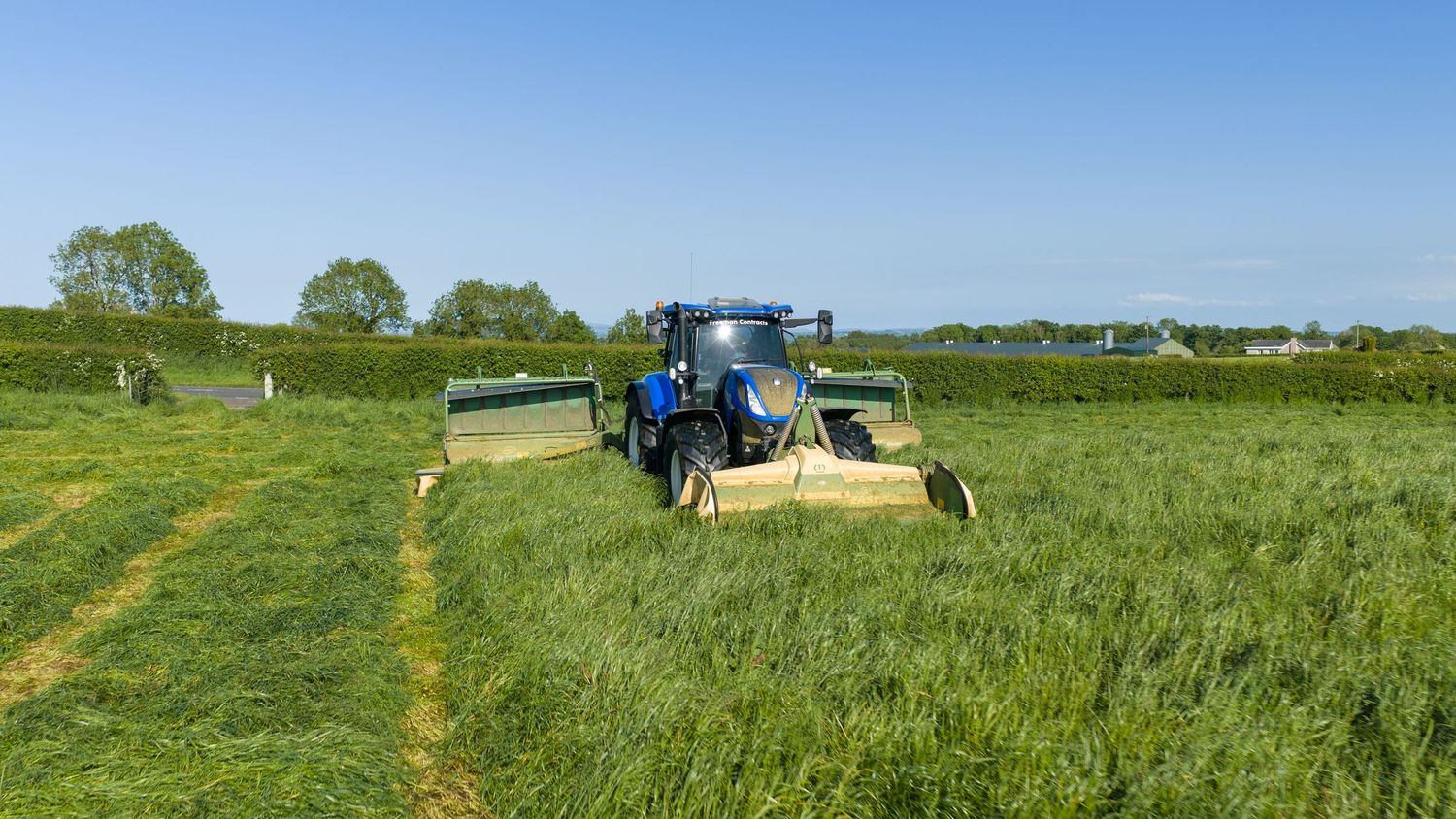Blue tractor mowing lush green grass field on a sunny day with trees in the background.