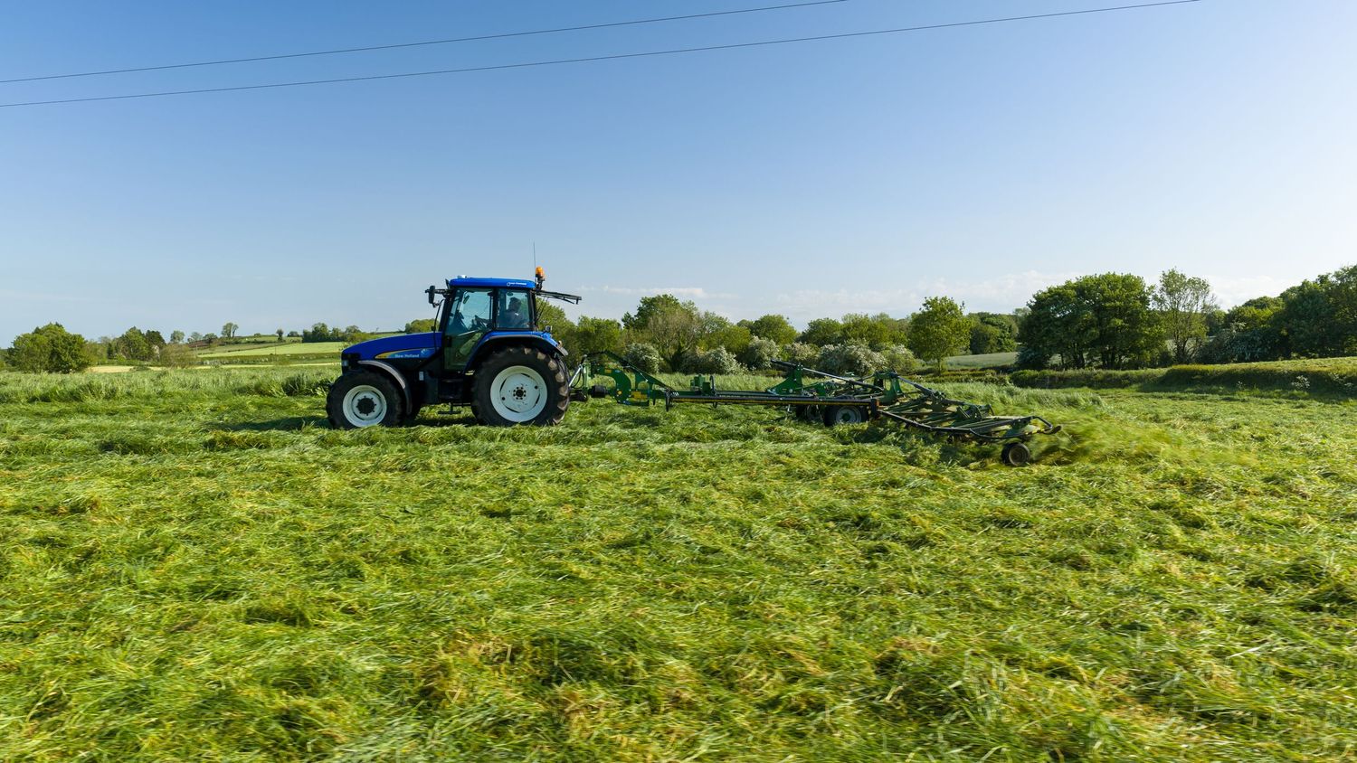 Blue tractor pulling green hay rake across lush farm field under clear blue sky on a sunny day.