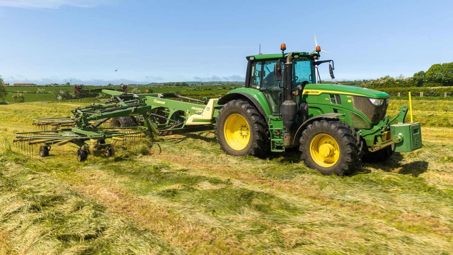 Green John Deere tractor raking cut hay in a lush field under a clear blue sky.