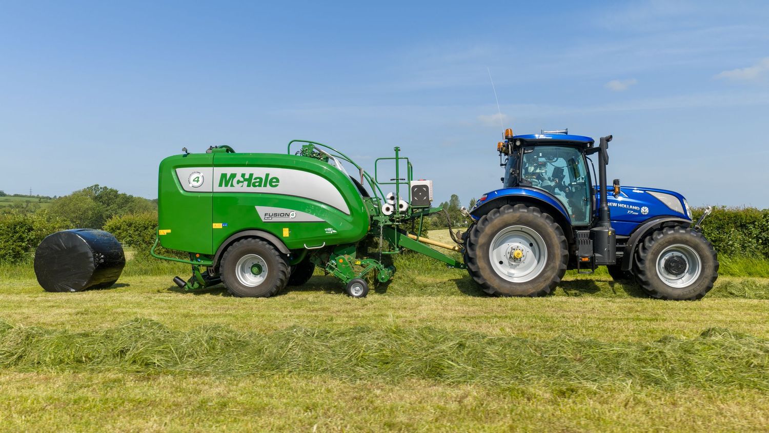 A blue New Holland tractor pulling a green McHale round baler in a sunny field during hay harvesting season.
