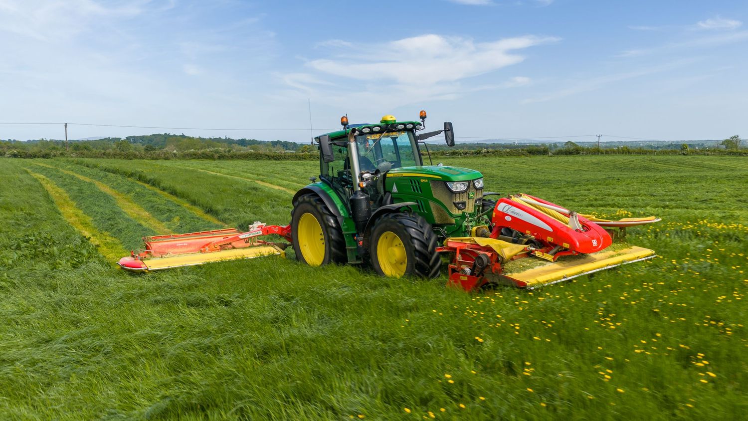 Green John Deere tractor with red mowers cutting lush green grass field on a sunny day.
