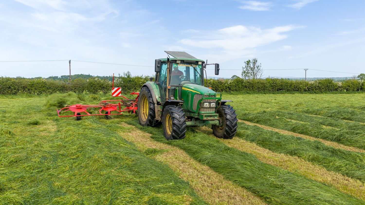Green John Deere tractor with red rake attachment cutting hay in a lush green field on a sunny day.